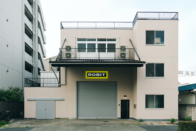 建物の全景写真。町工場らしい外観で、中央のシャッター上にはロゴマークの看板が取り付けられ、対比が意図されている。